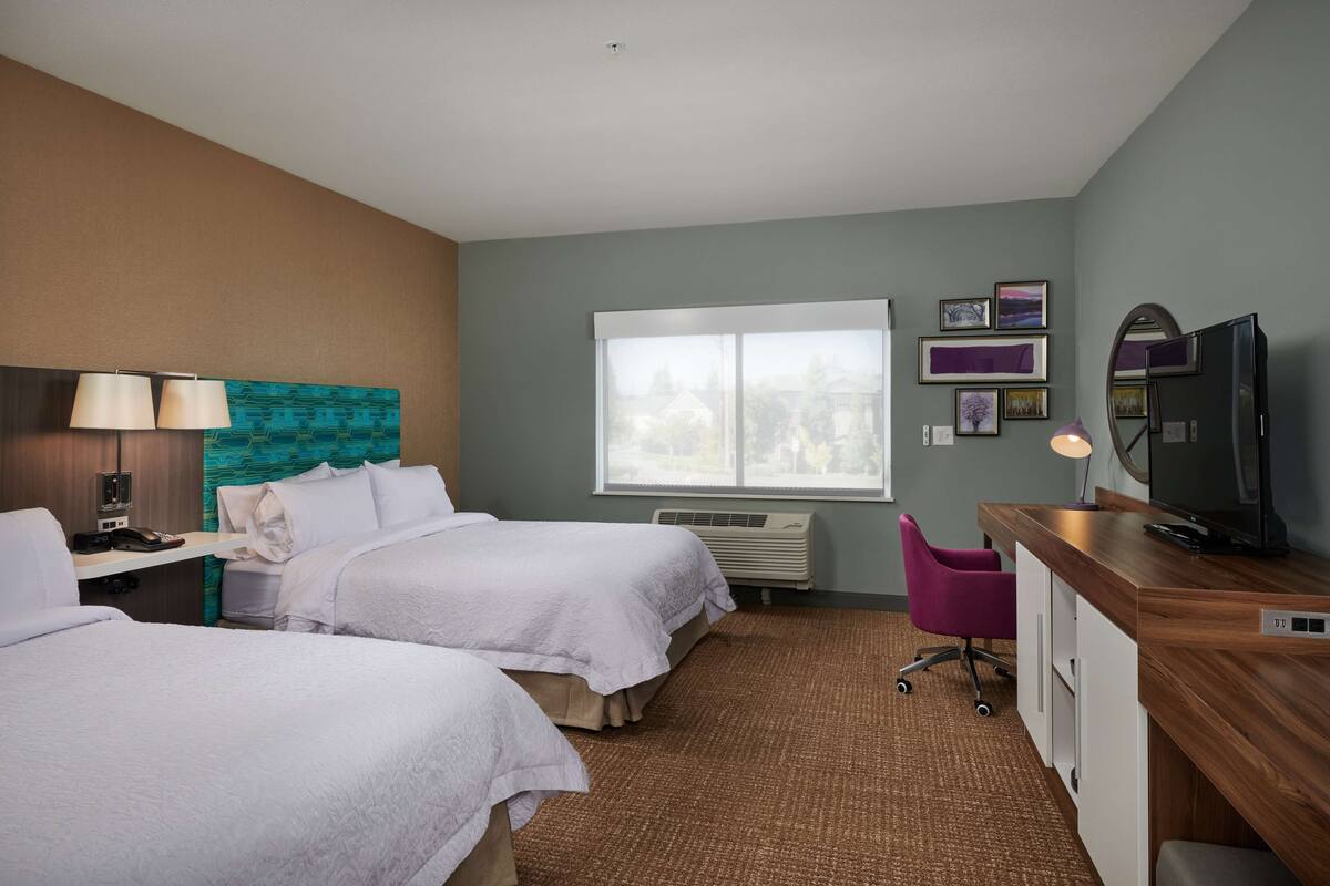 A hotel room with two beds, a desk, a chair, a TV, and a window with a view.