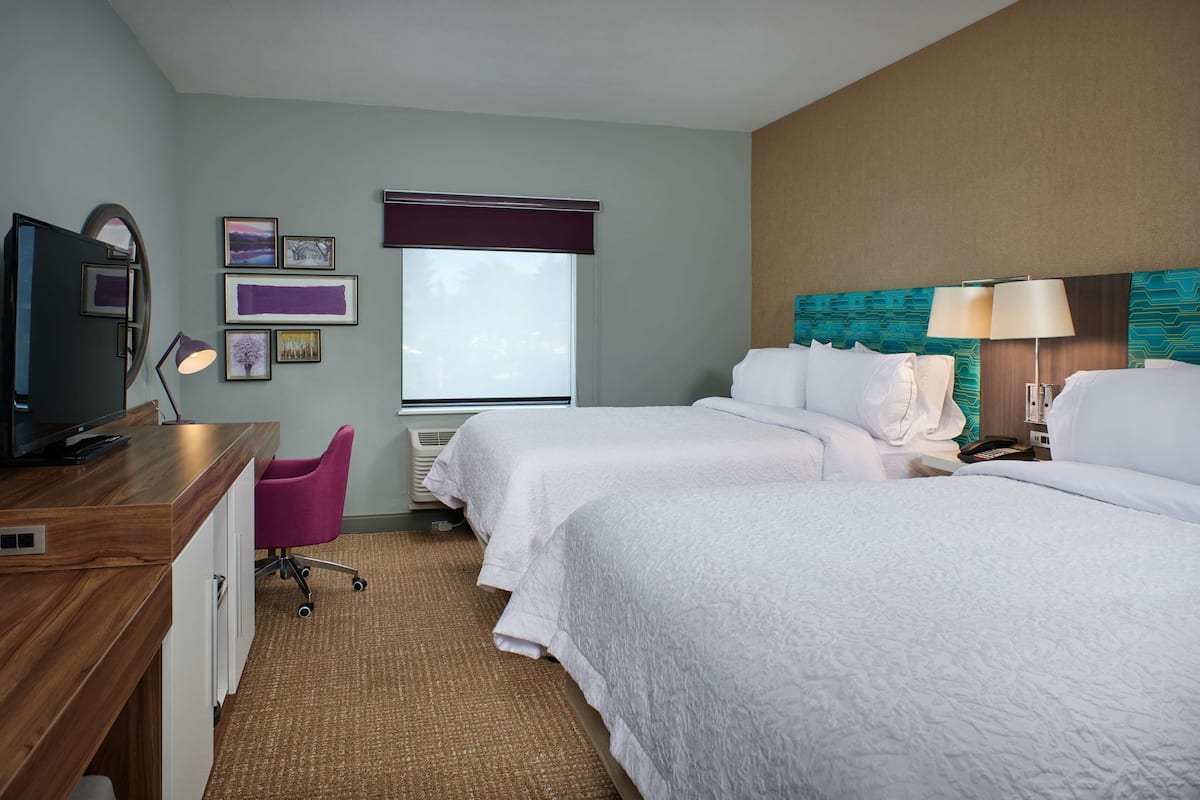 A hotel room with two beds, a desk with a computer, a chair, and a window with blinds.