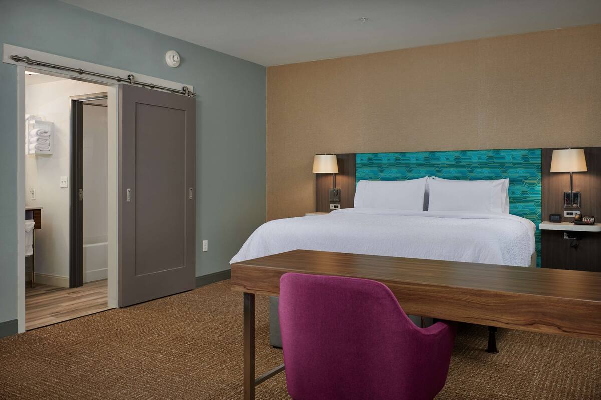 A hotel room with a bed, a desk, a chair, and a bathroom visible through an open door.