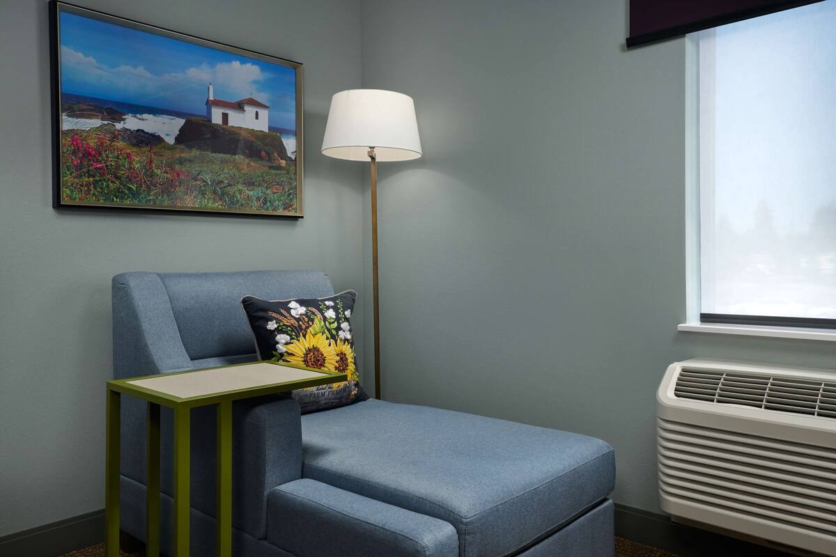 A room with a blue armchair, a small table, a floor lamp, a wall-mounted air conditioner, and a framed picture of a coastal scene.