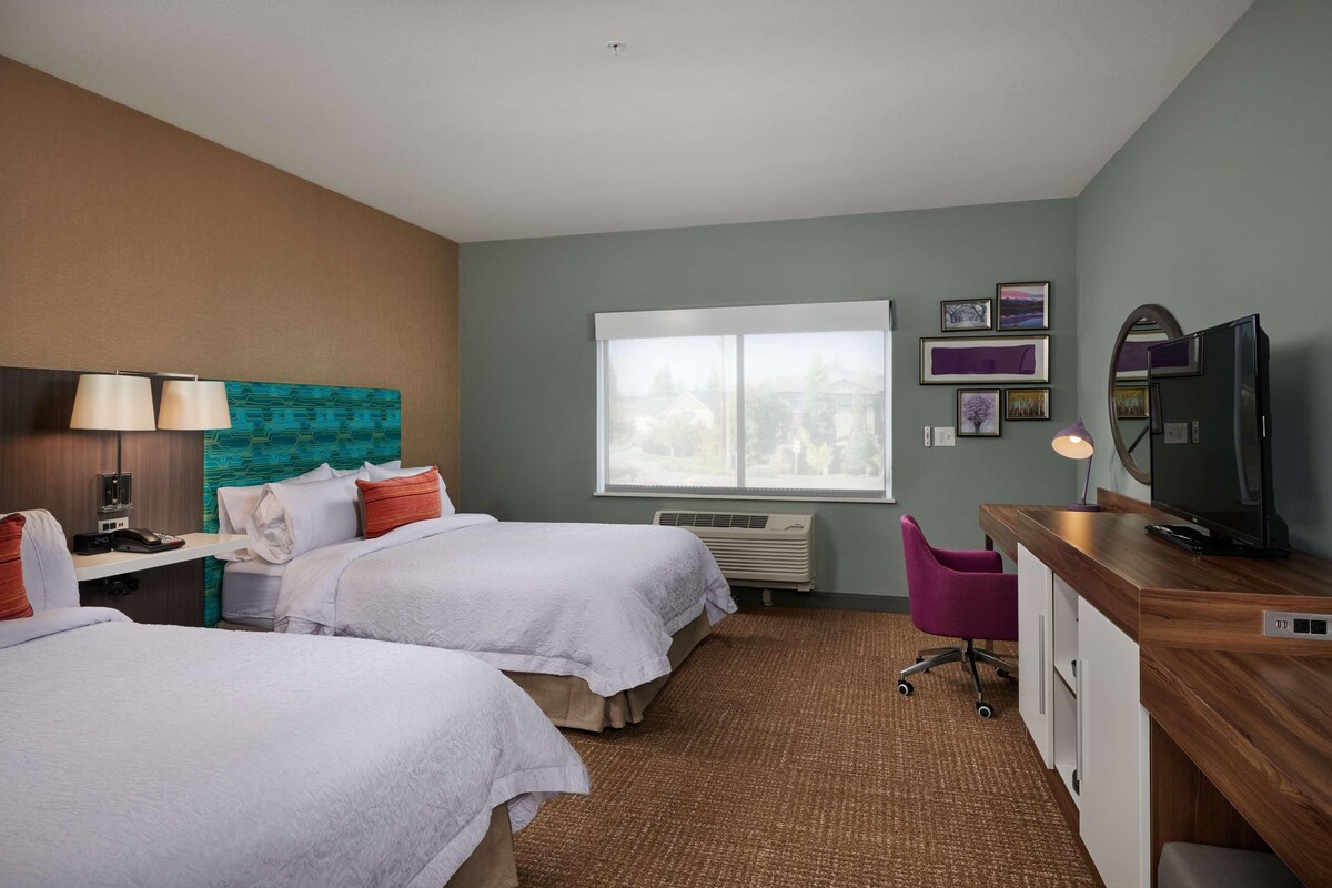 A hotel room with two beds, a desk with a computer, a TV, and a window with a view of the outside.