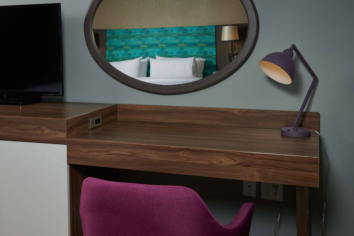 A hotel room with a wooden desk, a purple chair, a round mirror, and a bed with a teal patterned headboard.