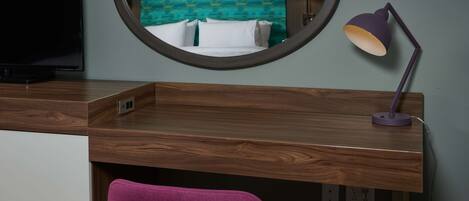 A hotel room with a wooden desk, a purple chair, a round mirror, and a bed with a teal patterned headboard.