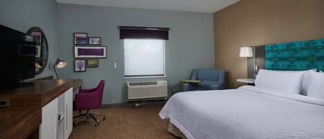 A hotel room with a bed, desk, chair, TV, air conditioning unit, and artwork on the walls.