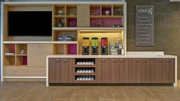 A modern hotel reception area with a large flat-screen TV, a 'Home2 Guarantee' sign, and a coffee station with decaf, gourmet, and hot cocoa options.