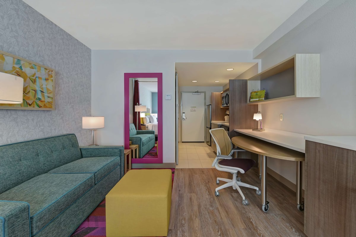 Et moderne hotelværelse med en grøn sofa, en gul skammel, et skrivebord med stol og et tekøkken.