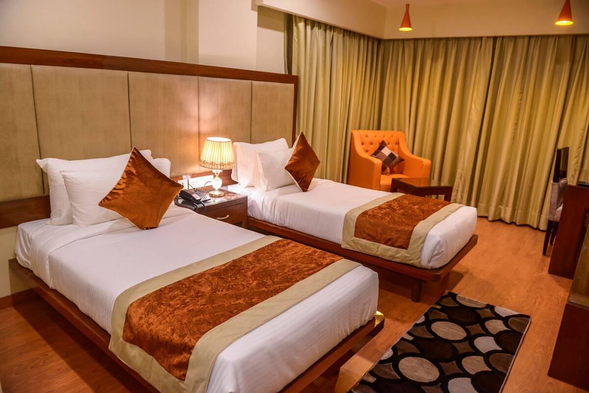A hotel room with two beds, a nightstand, a lamp, a chair, and a large window with curtains.