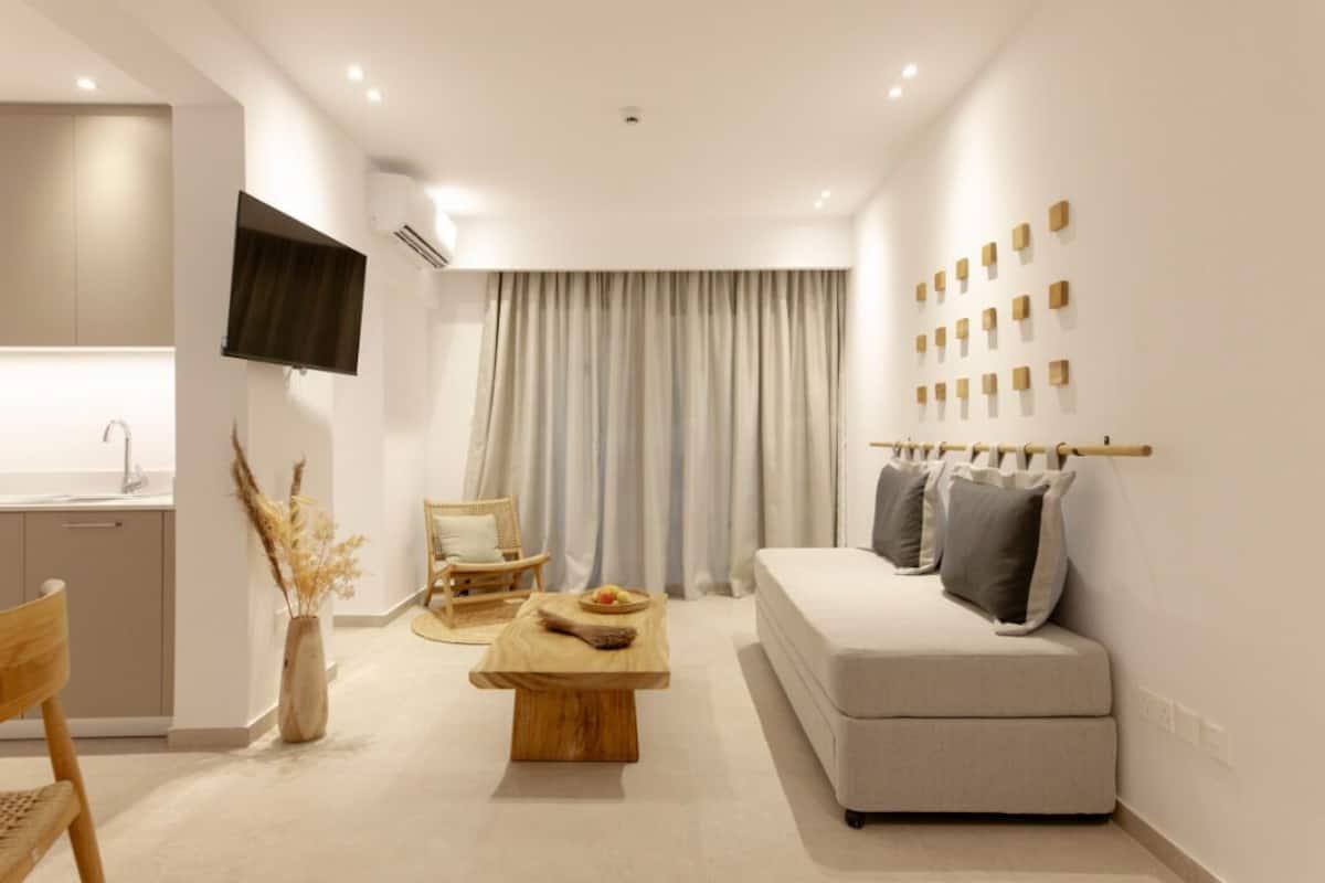 A modern living room with a sofa, a wooden coffee table, a dining table, and a chair. There is a kitchen area with a sink and cabinets.