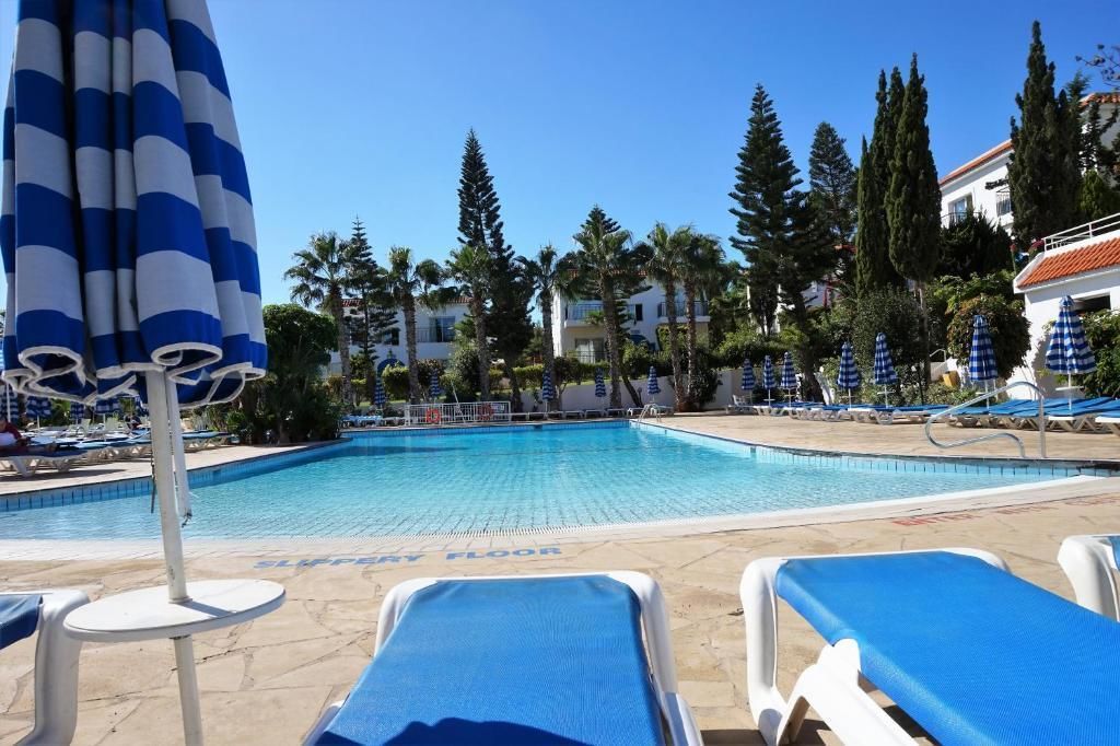 Un espace piscine agrémenté de chaises longues et de parasols, entouré de palmiers et d’immeubles.
