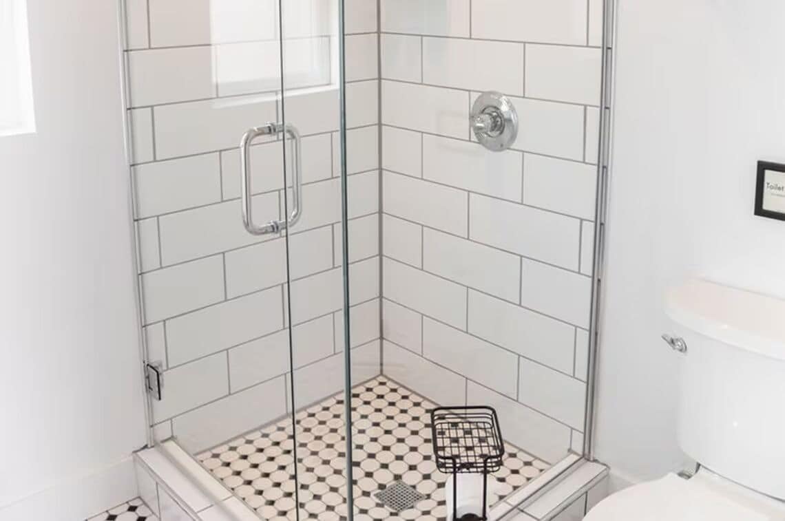 A corner bathroom with a glass shower enclosure, white tiled walls, and a checkered floor pattern.