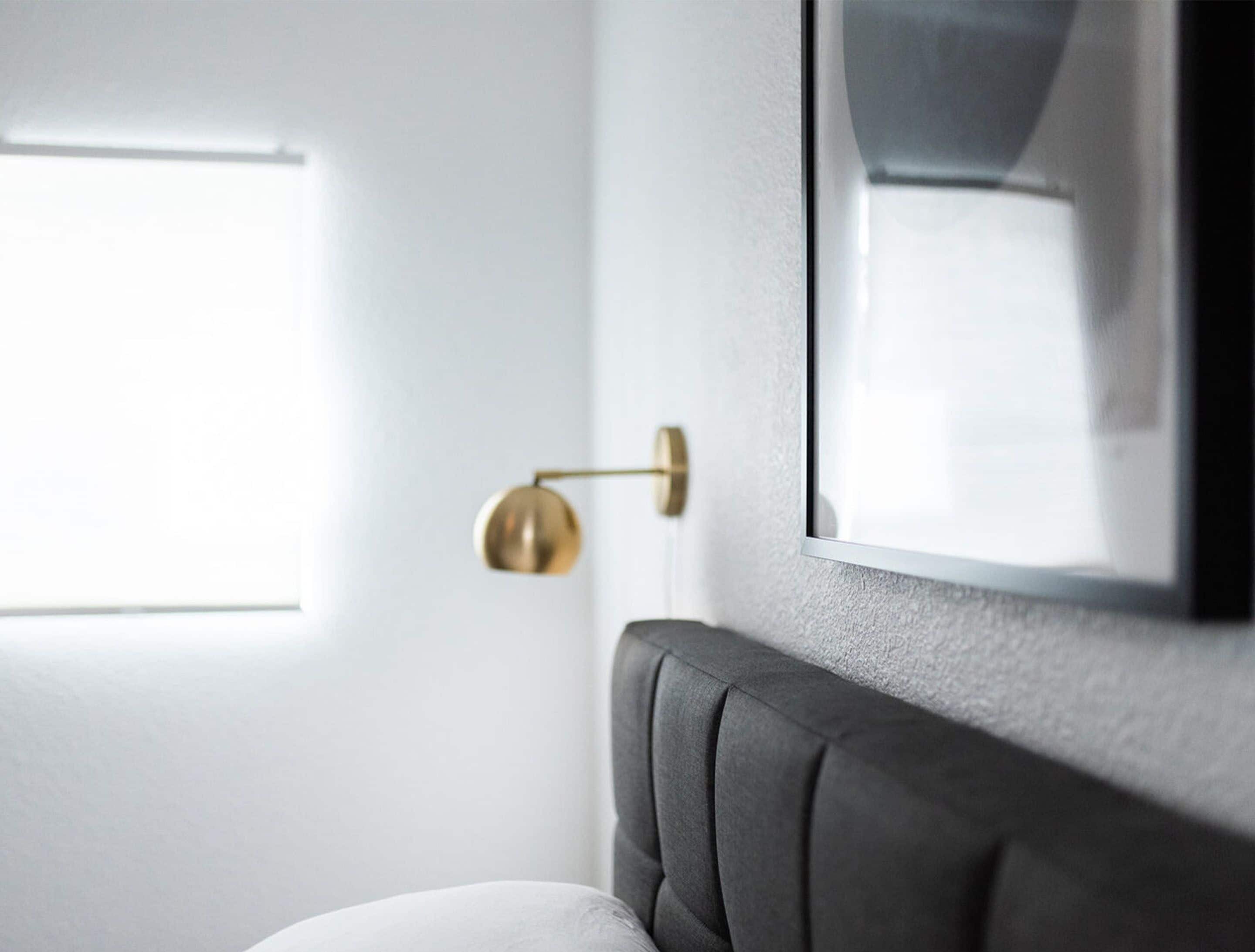 A modern room with a grey upholstered headboard, a wall-mounted round gold lamp, and a framed mirror reflecting a window.