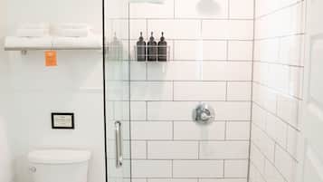 A modern bathroom with a glass-enclosed shower, white tiles, a toilet, and a shelf with folded towels.