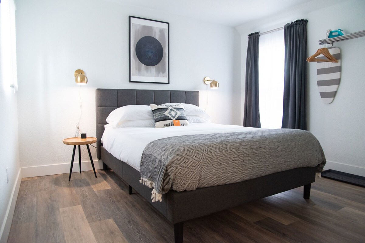 A modern bedroom with a grey upholstered bed, a small bedside table, a wall-mounted lamp, and a framed abstract artwork.