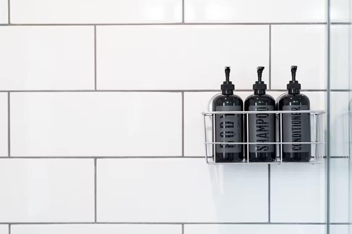 A bathroom with a white tiled wall and a metal shelf holding three black bottles labeled "SHAMPOO" and "CONDITIONER".