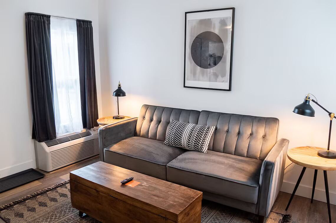 A modern living room with a leather sofa, a wooden coffee table, a side table with a lamp, and a large window with curtains.