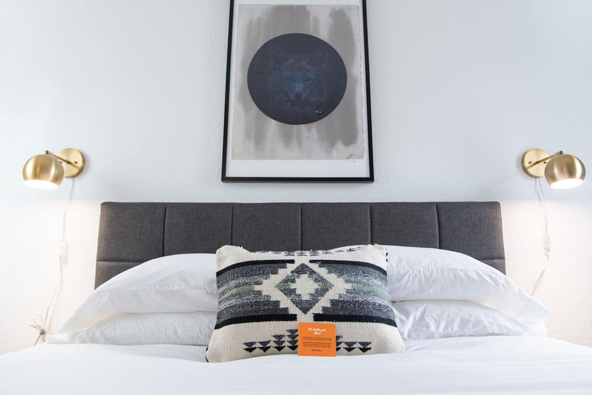 A neatly made bed with white and patterned pillows, a framed abstract artwork above, and two wall-mounted lamps.