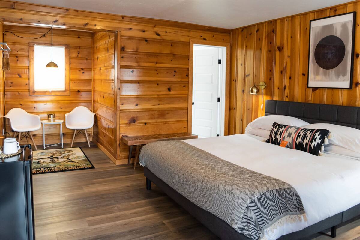 A wooden-paneled room with a bed, two white chairs, a small table, and a door.
