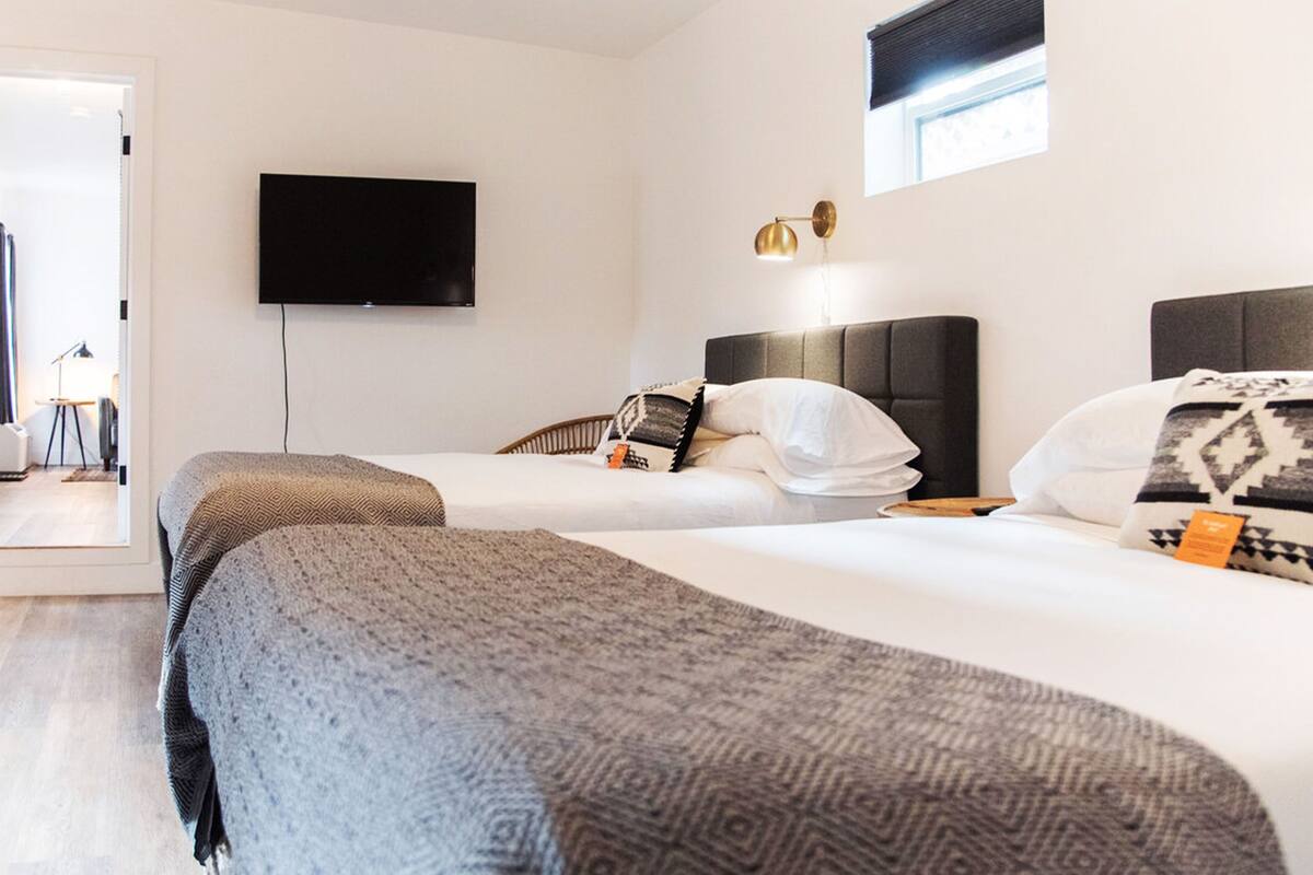 A hotel room with two beds, a flat-screen TV mounted on the wall, a window with a blind, and a bedside lamp.