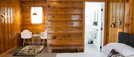 A wooden room with a bed, a desk, two chairs, a rug, and a bathroom visible through an open door.