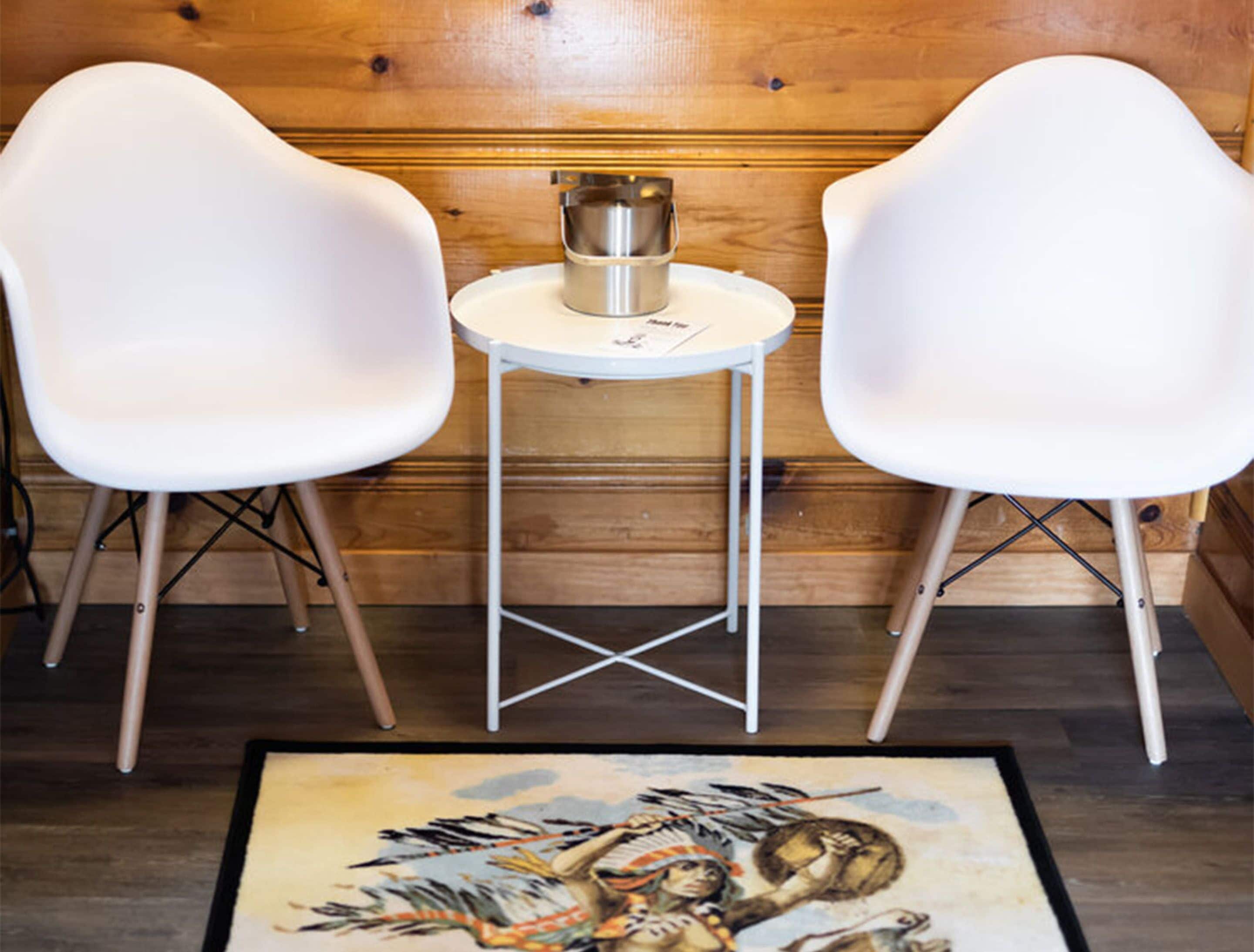 Deux chaises blanches et une petite table surmontée d’un pichet en métal, dans une pièce aux murs lambrissés et ornée d’un tapis à motifs.