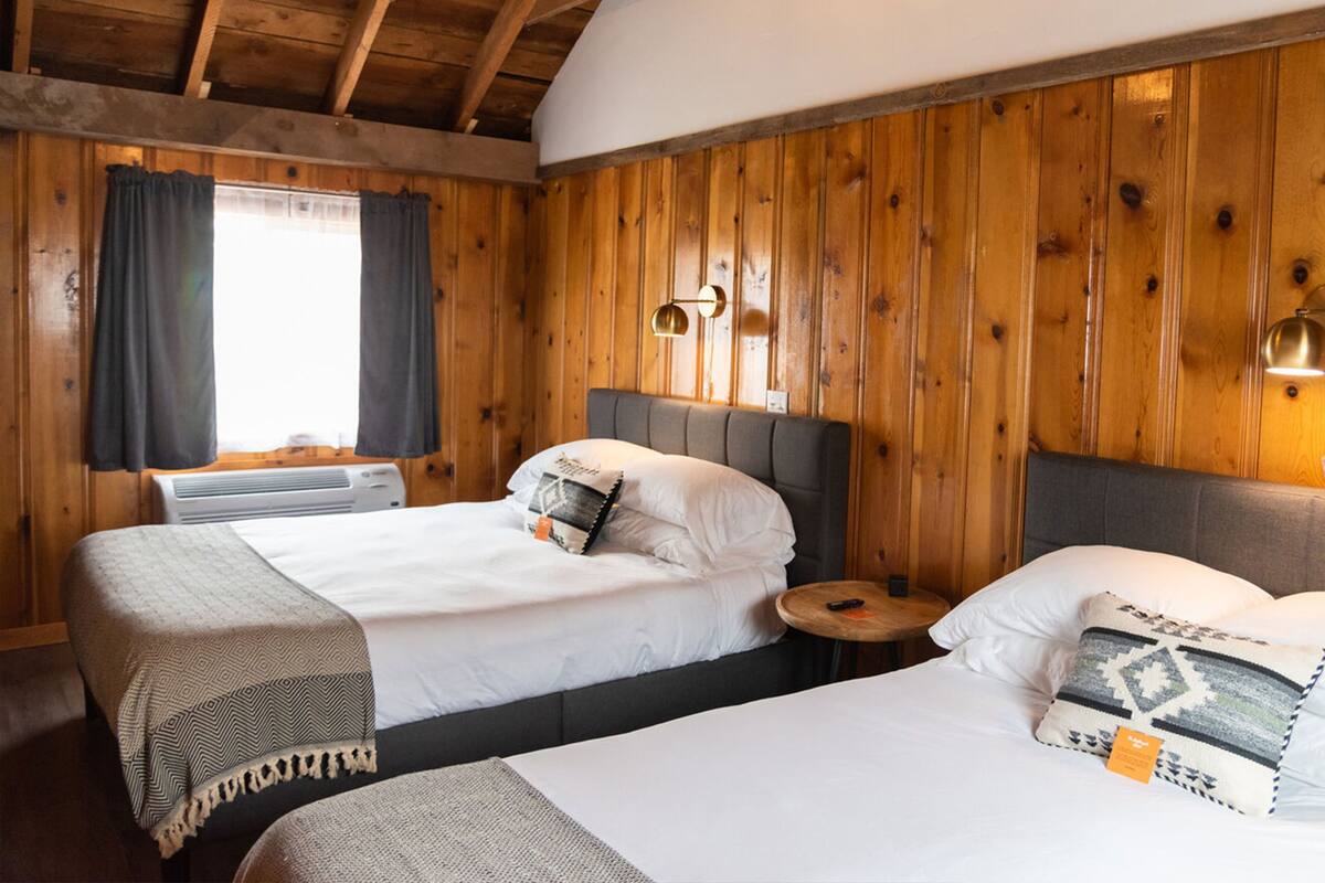 A room with two beds, wooden walls, a window with curtains, and a small round table with a lamp.