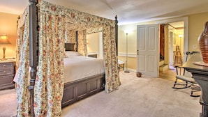 A four-poster canopy bed with floral curtains, a bedside table, a lamp, and a rocking chair in a room with beige walls and carpet.