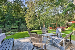 Relax in a serene outdoor space surrounded by lush greenery and a small pond, with a stone table and Adirondack chairs.