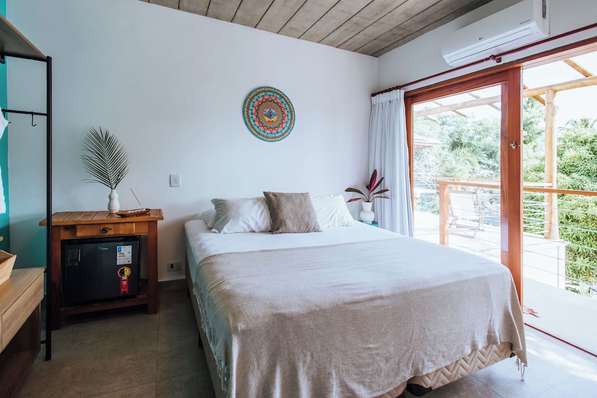 Un dormitorio con cama, una mesita de madera, aire acondicionado de pared y vista a la vegetación a través de una puerta corrediza de vidrio.
