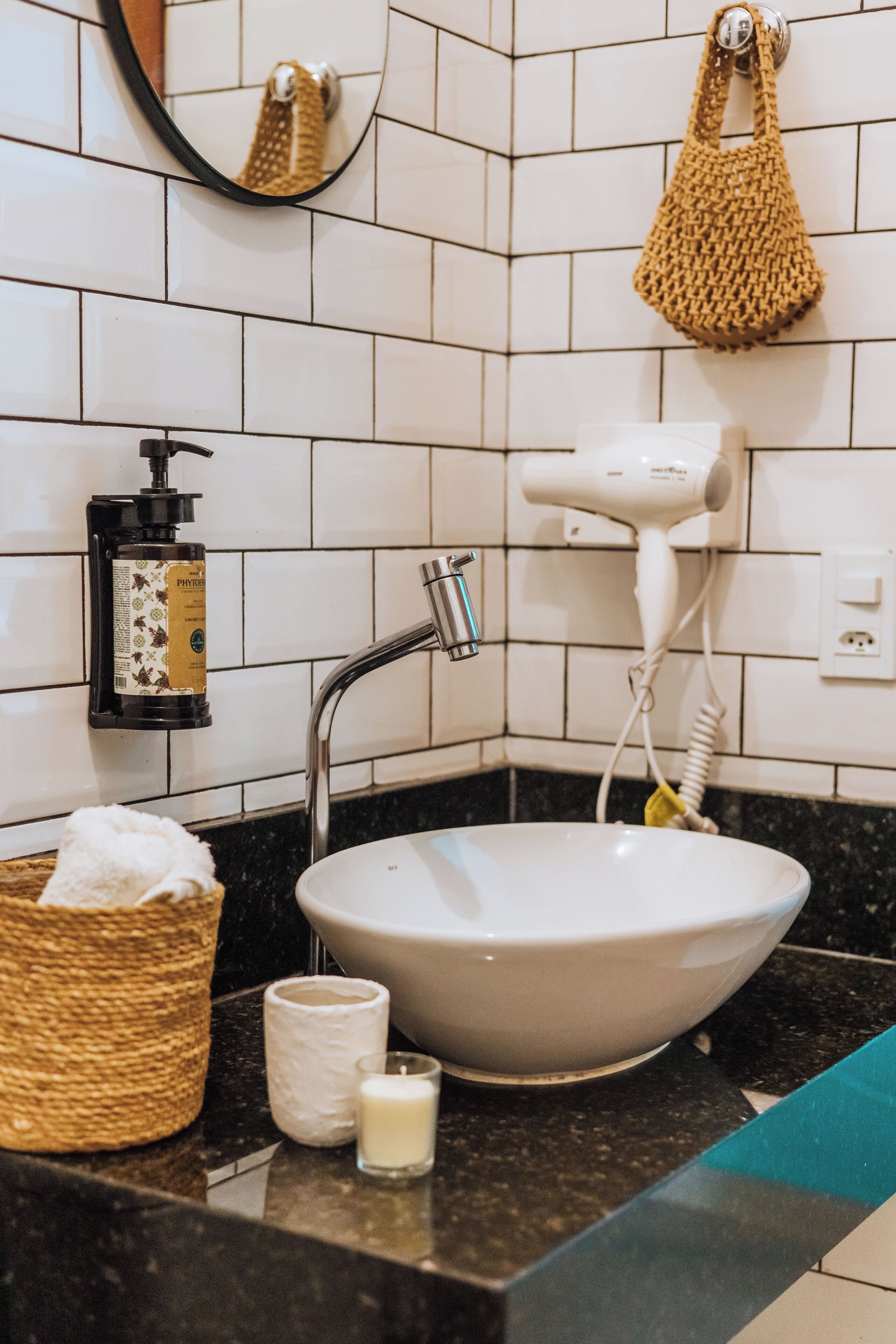 A bathroom with a white sink, a hairdryer, a bottle of shampoo, a candle, and a woven bag.