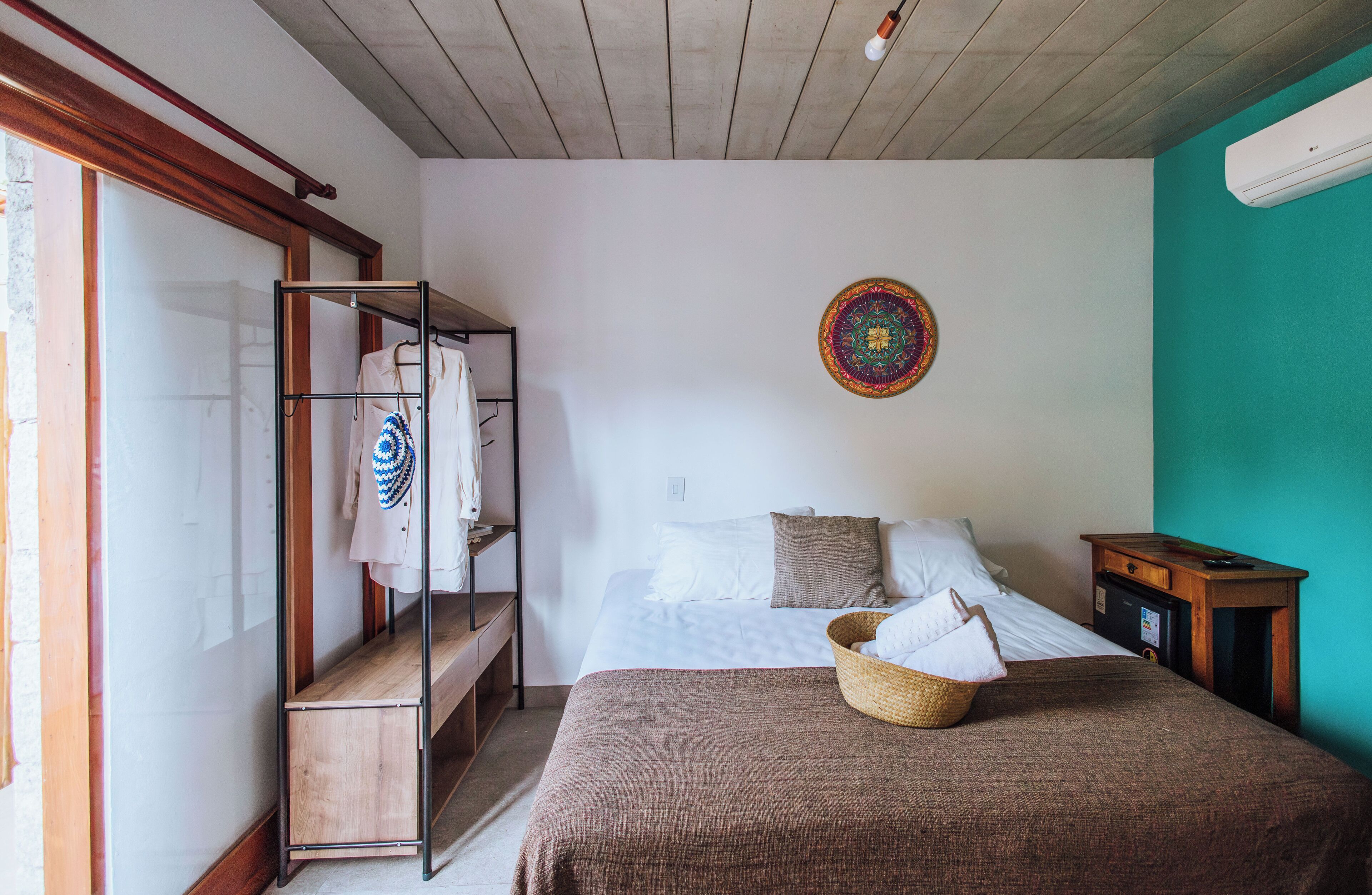 Ein Schlafzimmer mit einem Bett, einem Holzregal mit Kleidung, einem farbenfrohen Wandbehang und einer türkisblauen Akzentwand.