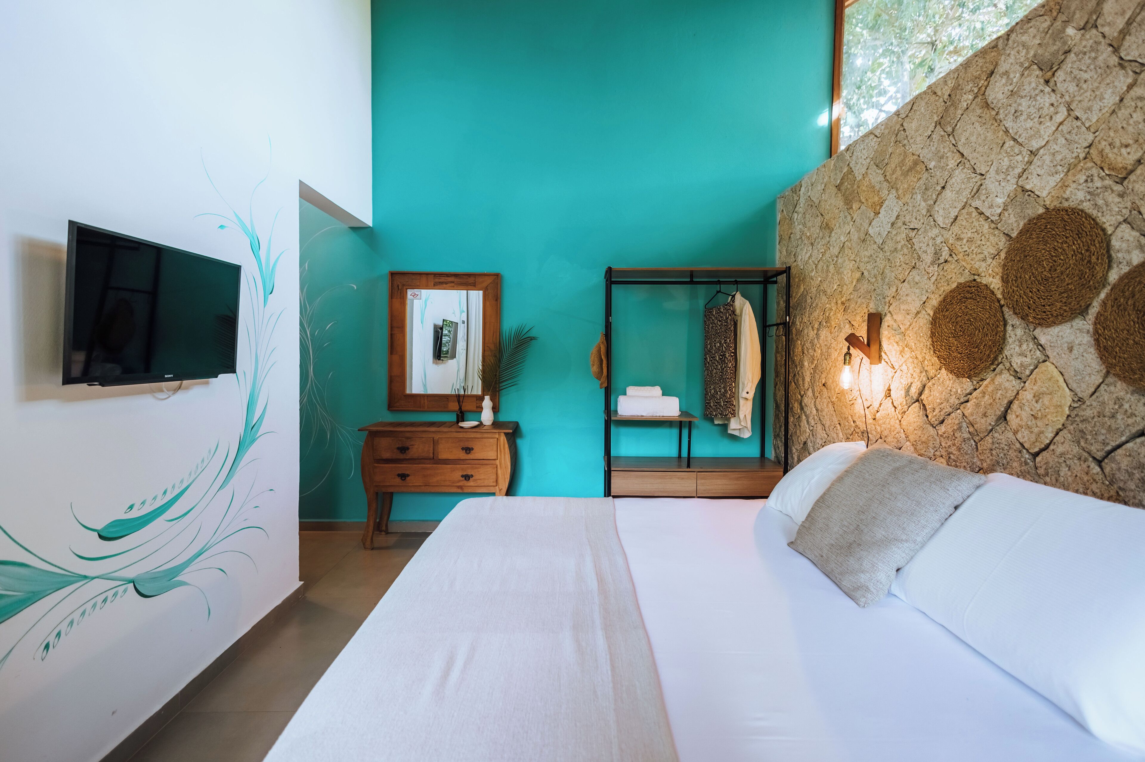 Un dormitorio con cama, televisión, cómoda de madera, espejo y pared de piedra con decoraciones tejidas.