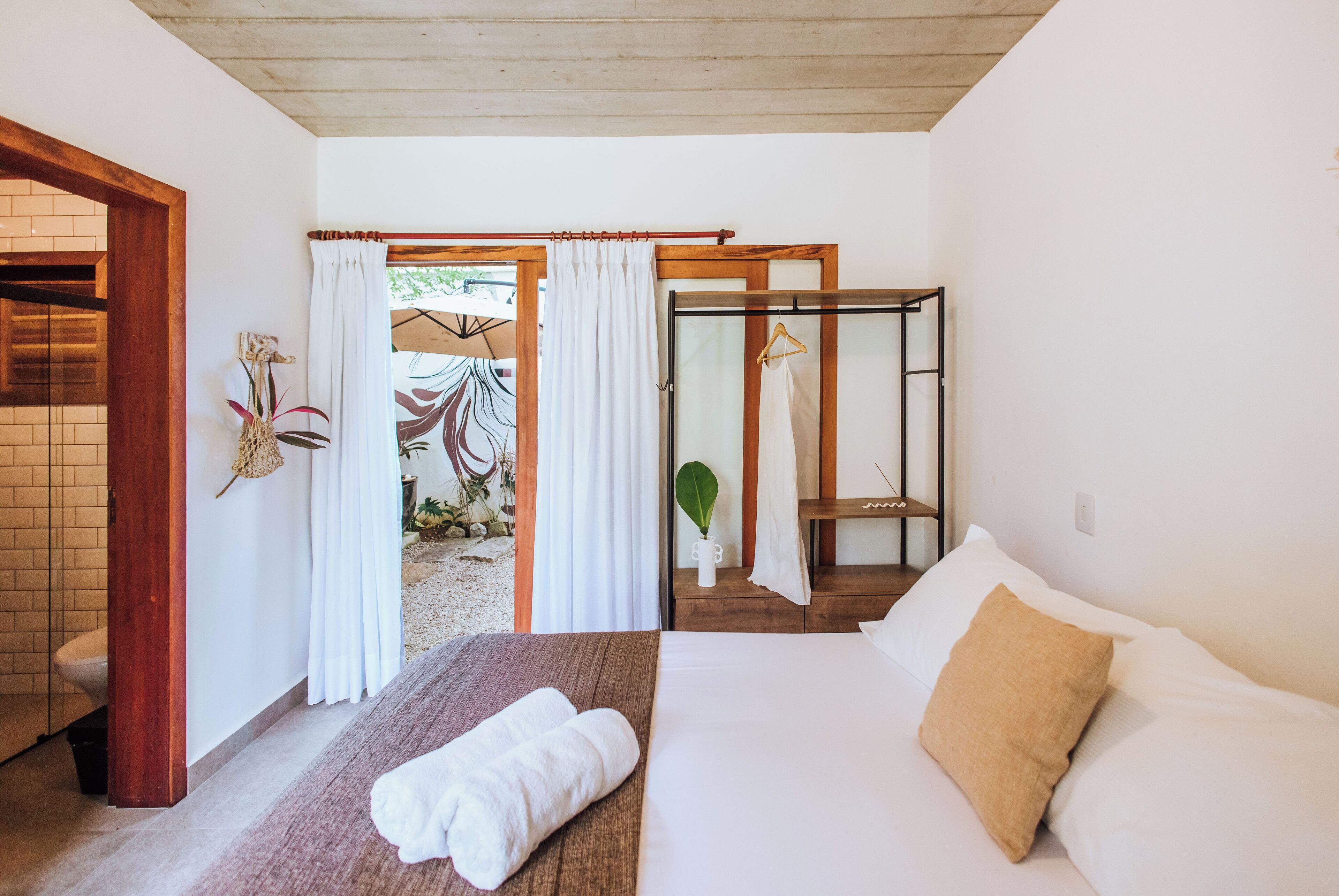 Un dormitorio con cama, un banco de madera, una mesita con una planta y un baño visible a través de una puerta abierta.