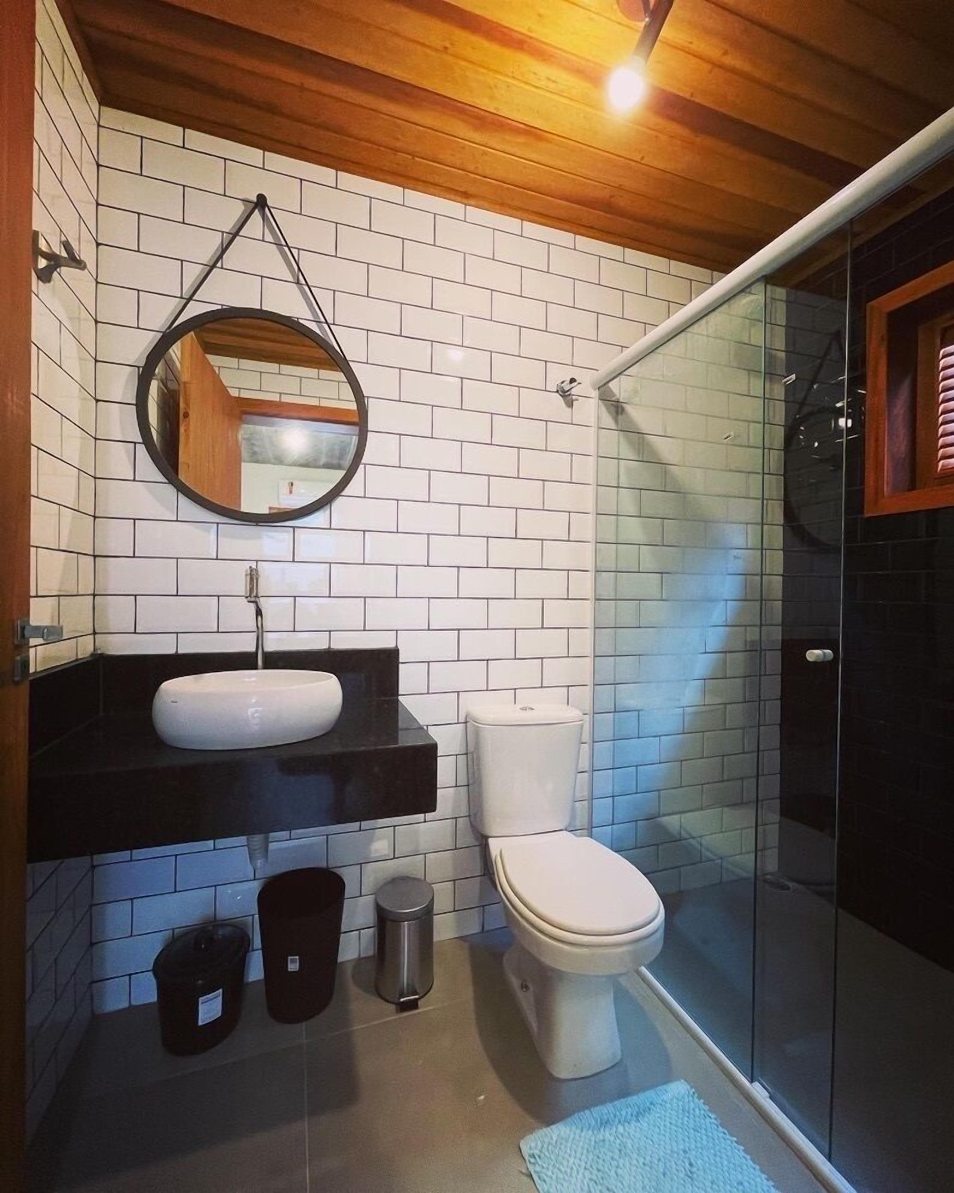 A modern bathroom with a white-tiled wall, a wooden ceiling, a round mirror, a black countertop, a white toilet, and a glass shower enclosure.