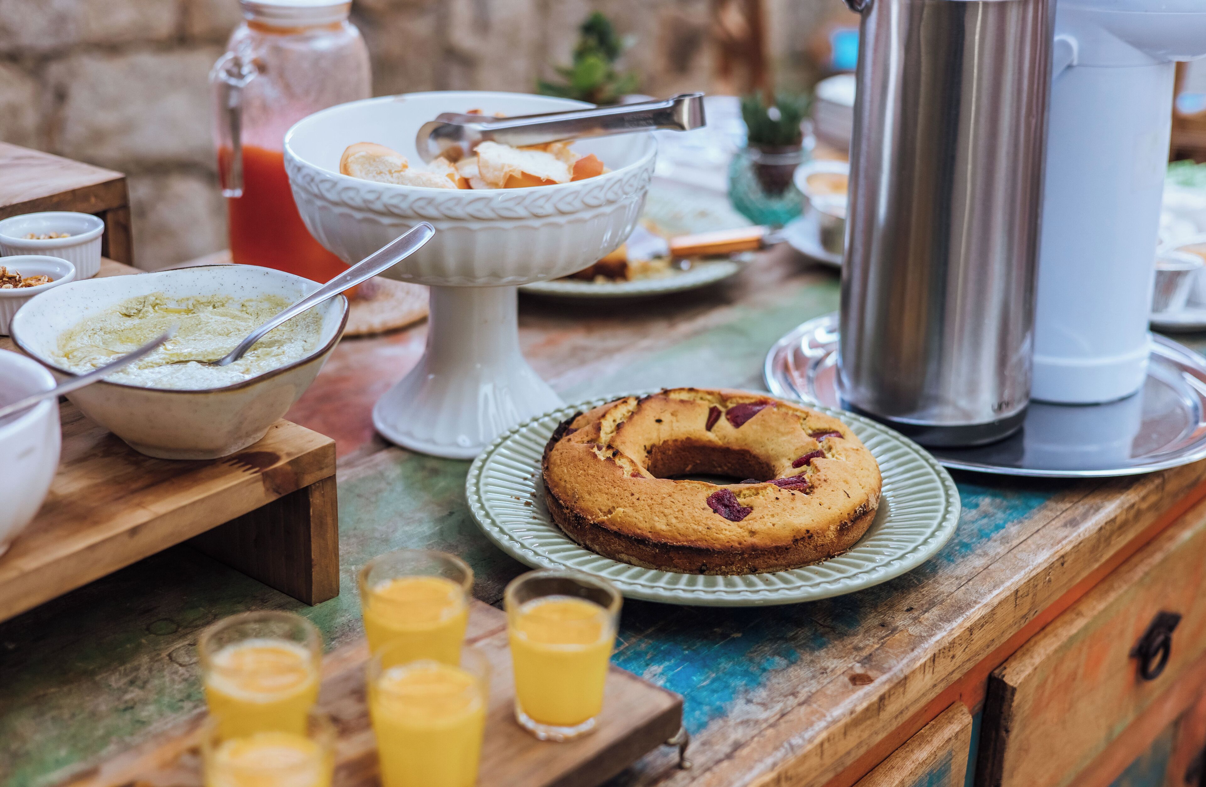 Ein gedeckter Frühstückstisch mit verschiedenen Speisen, darunter ein Kuchen auf einem Teller, Schüsseln und Gläser Orangensaft.