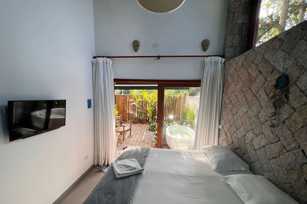 Un dormitorio con cama, televisión, pared de piedra y vista a un jardín.