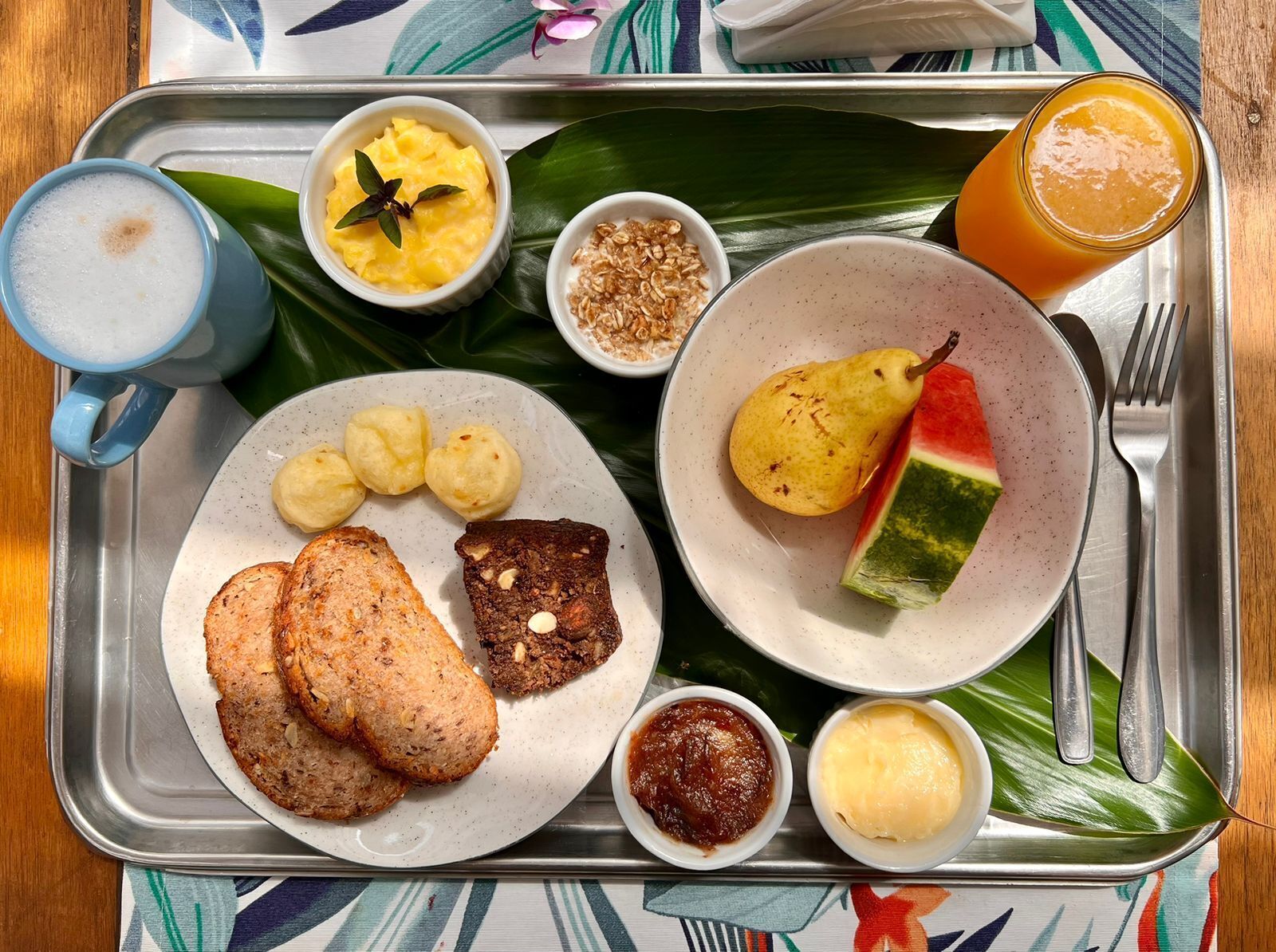 Un plateau contenant divers éléments pour le petit-déjeuner, notamment des toasts, des œufs brouillés, une poire, une tranche de pastèque et plusieurs petits ramequins de condiments.