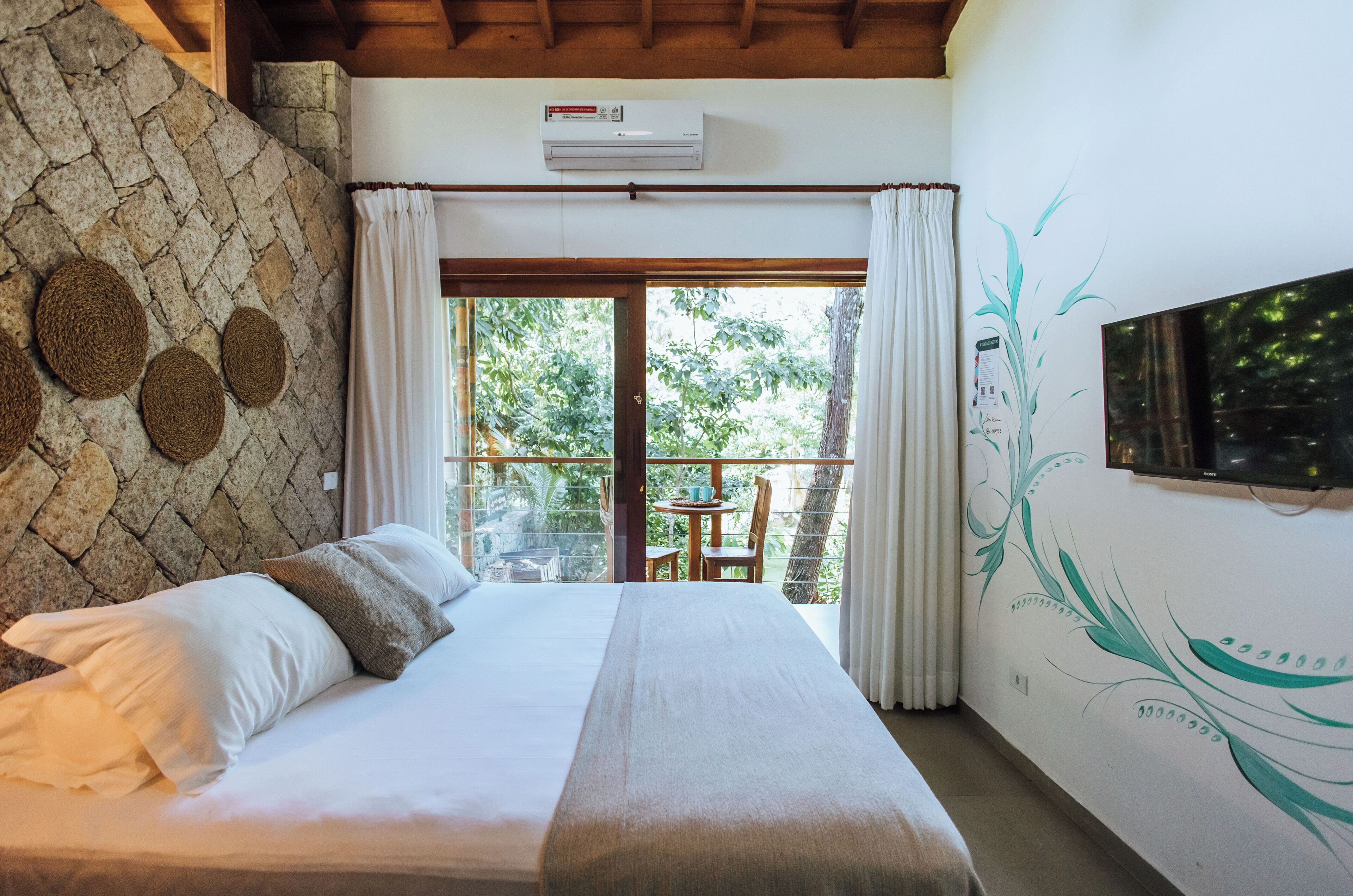 Una camera da letto con un letto, parete in pietra, condizionatore d'aria e vista sul verde attraverso una porta in vetro.
