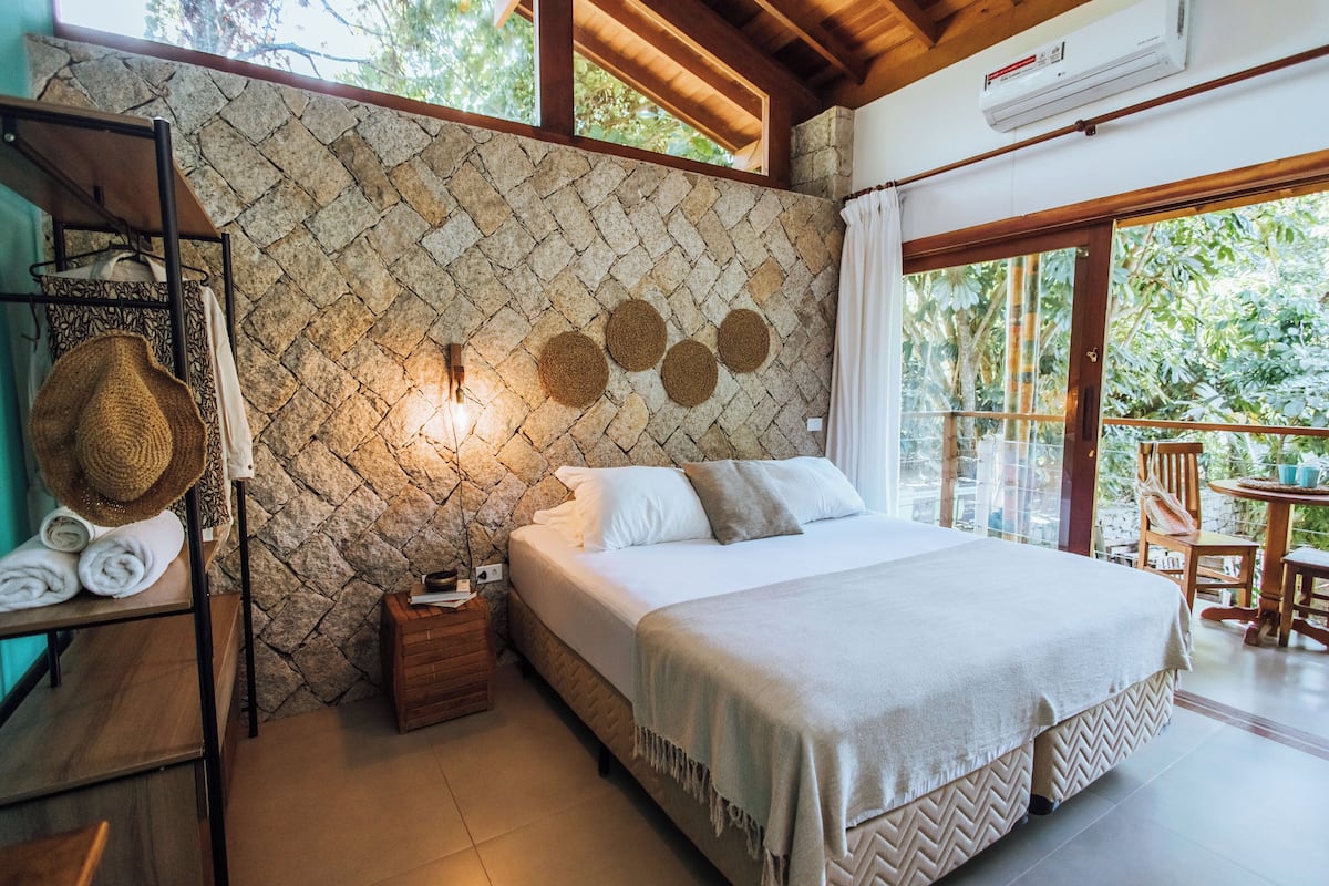 Un dormitorio con pared de piedra, una cama con colcha blanca, una mesita de noche de madera, una cesta de mimbre y vistas a la vegetación exterior.
