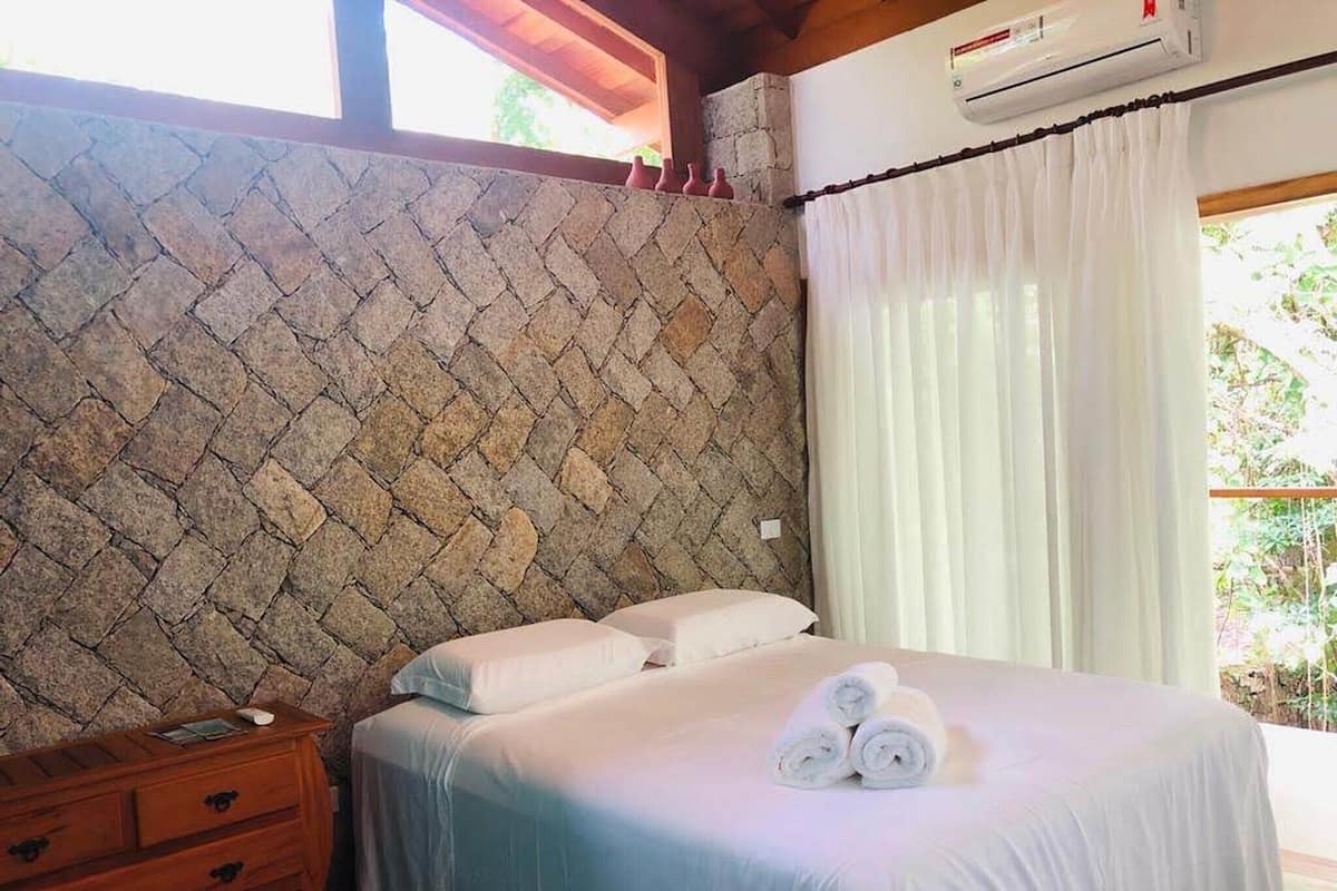 Un dormitorio con pared de piedra, una cama con ropa blanca, una mesita de noche de madera y una ventana con cortinas blancas.