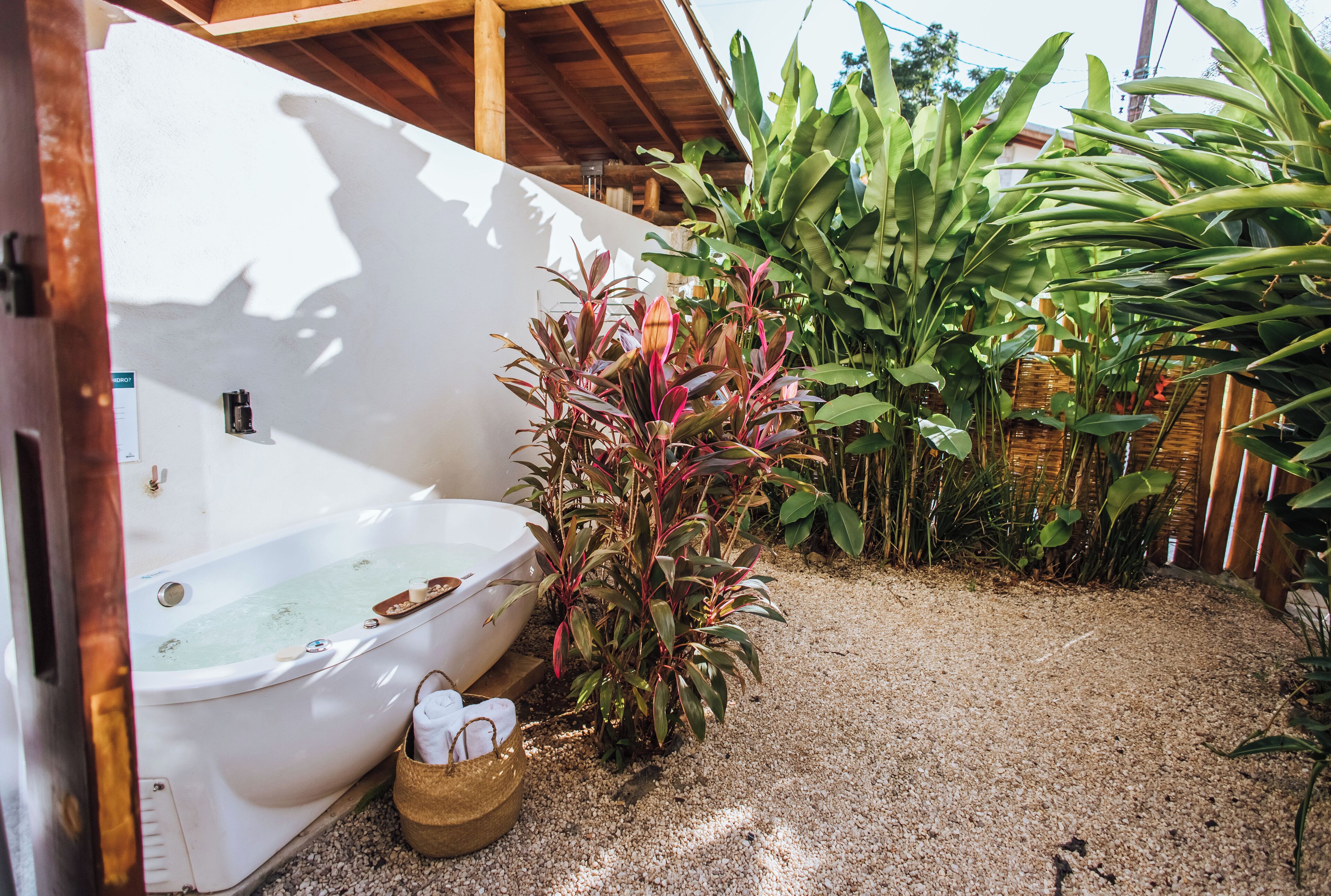 Une baignoire blanche avec un banc en bois, entourée de plantes et posée sur un sol de gravier.