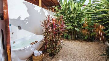 A white bathtub with a wooden bench, surrounded by plants and a gravel floor.
