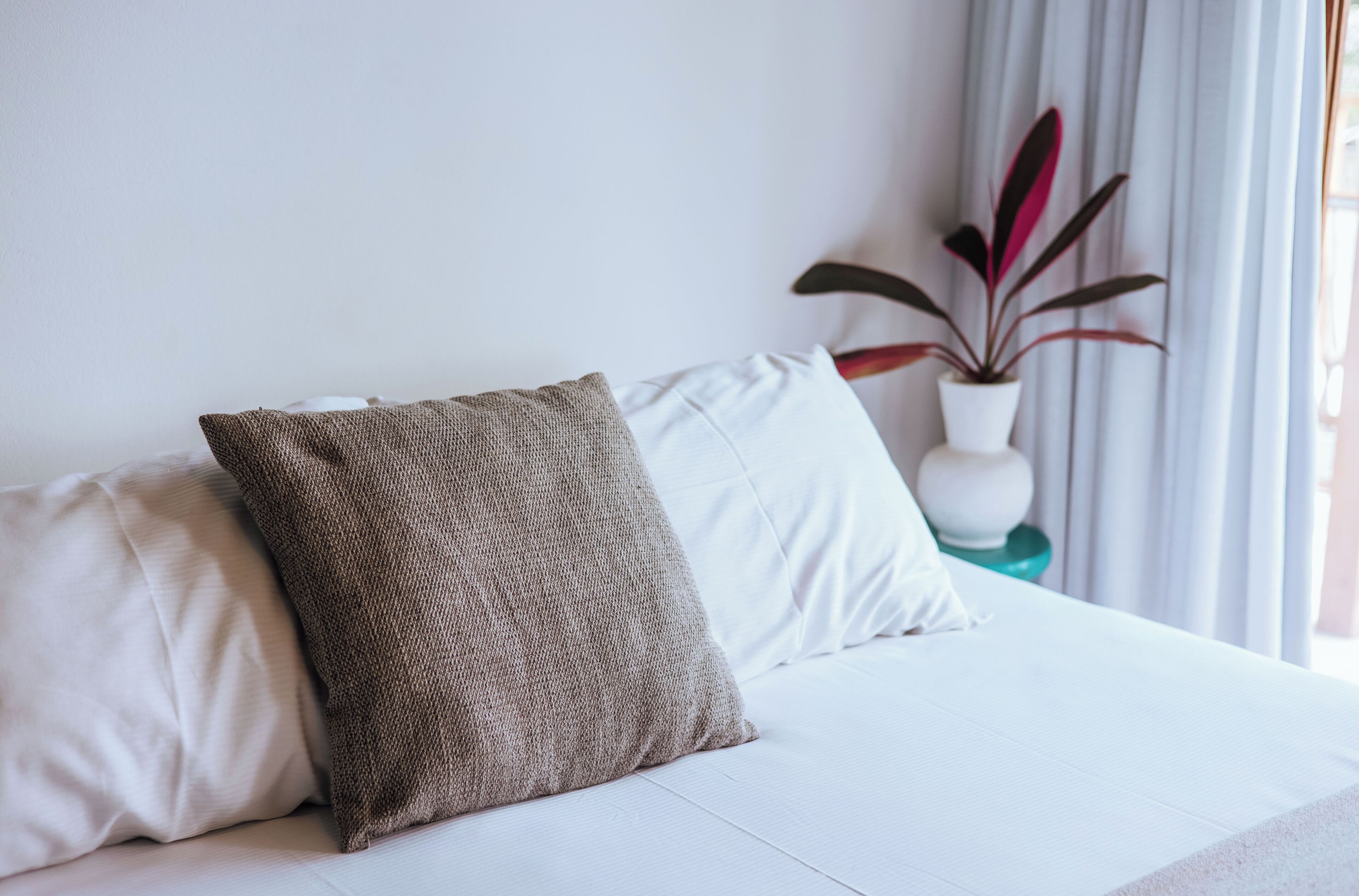 A bed with white and beige pillows, a white vase with a plant, and a window with curtains.