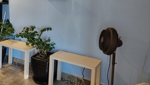 A room with a light blue wall, a framed painting of horses, a white shelf, a potted plant, and a standing fan. - Travel Inn Delaware (Delaware)