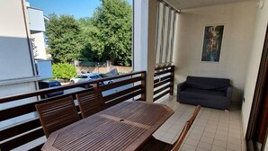 Varanda com mesa e cadeiras de madeira, um sofá cinza e vista para carros estacionados e árvores.