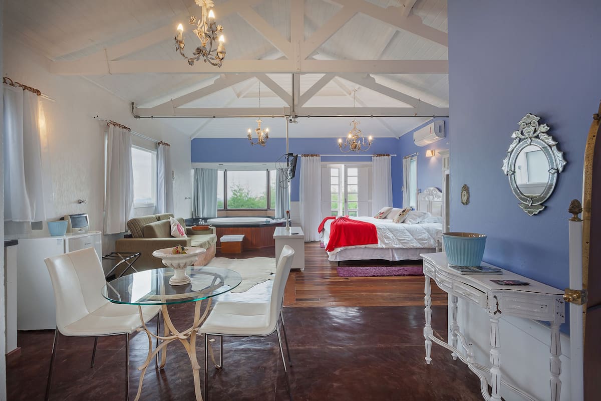 A spacious room with a glass dining table, white chairs, a bed with a red blanket, and a large mirror on the wall.