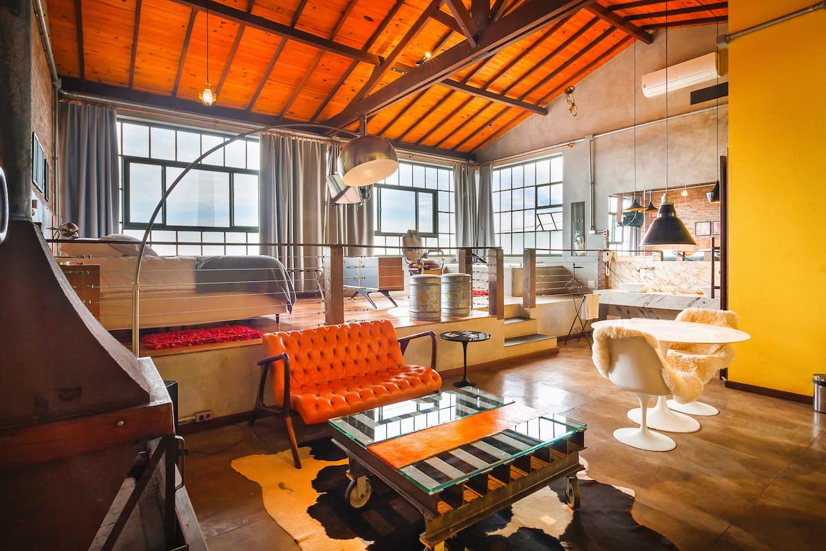 A spacious living area with a high ceiling, large windows, a sofa, a glass coffee table, and a dining table with chairs.