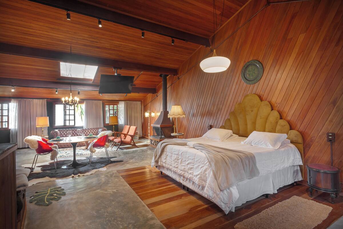 A room with a large bed, wooden ceiling, a dining table with chairs, and a fireplace.