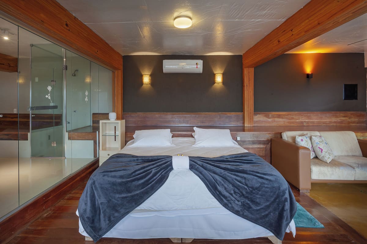 A bedroom with a bed, a sofa, a glass shower enclosure, and an air conditioning unit.