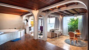 Um quarto espaçoso com teto de madeira, uma janela grande com cortinas, uma área de jantar com mesa e cadeiras, e um banheiro com banheira e pia.