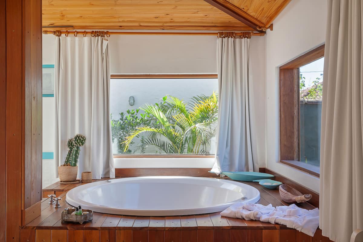 A bathroom with a large bathtub, a window with curtains, and a view of greenery outside.